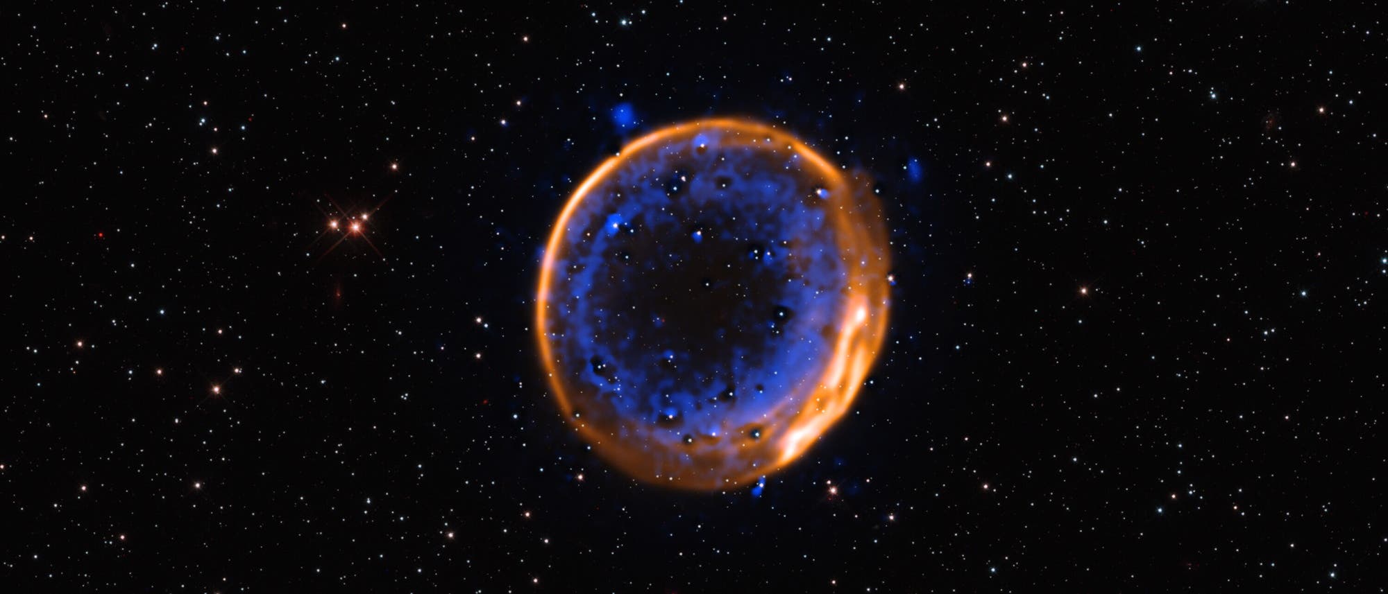 Diese Aufnahme zeigt die farbenfrohen Überreste eines einstigen Weißen Zwergs: Der Supernova-Überrest SNR 0509 ist rund 160 000 Lichtjahre von der Erde entfernt und befindet sich in der Großen Magellanschen Wolke, einer Satellitengalaxie der Milchstraße. Dieser Supernova-Überrest ist der erste sichtbare Beweis dafür, dass Supernovae vom Typ Ia durch eine zweifache Explosion entstehen können: Darauf weisen die beiden konzentrischen und separaten Schalen an Kalzium in dieser Aufnahme hin, die hier in Blau dargestellt sind. Demnach ist einst zunächst die Hülle des Weißen Zwerges explodiert, was eine zweite Explosion seines Kerns ausgelöst hat.
Diese Aufnahme zeigt die farbenfrohen Überreste eines einstigen Weißen Zwergs: Der Supernova-Überrest SNR 0509 ist rund 160 000 Lichtjahre von der Erde entfernt und befindet sich in der Großen Magellanschen Wolke, einer Satellitengalaxie der Milchstraße. Dieser Supernova-Überrest ist der erste sichtbare Beweis dafür, dass Supernovae vom Typ Ia durch eine zweifache Explosion entstehen können: Darauf weisen die beiden konzentrischen und separaten Schalen an Kalzium in dieser Aufnahme hin, die hier in Blau dargestellt sind. Demnach ist einst zunächst die Hülle des Weißen Zwerges explodiert, was eine zweite Explosion seines Kerns ausgelöst hat.