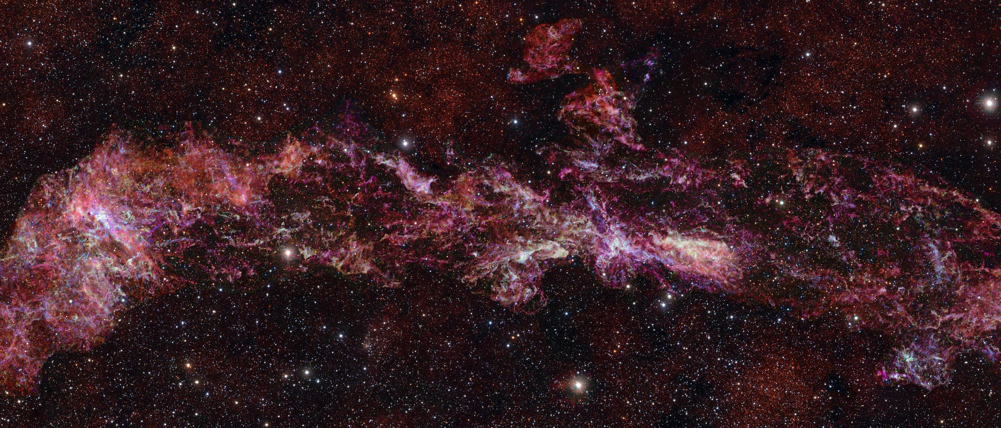 Ein farbenpr&auml;chtiges astronomisches Bild zeigt den Kernbereich unseres Milchstra&szlig;ensystems. Die Aufnahme zeigt komplexe, filamentartige Strukturen in leuchtenden Rot- und Pinkt&ouml;nen, die sich vor einem Hintergrund aus unz&auml;hligen Sternen erstrecken. Der Nebel erscheint wie ein leuchtendes Band, das sich &uuml;ber das Bild zieht, und hebt die dynamische und chaotische Natur solcher kosmischen Ereignisse hervor.