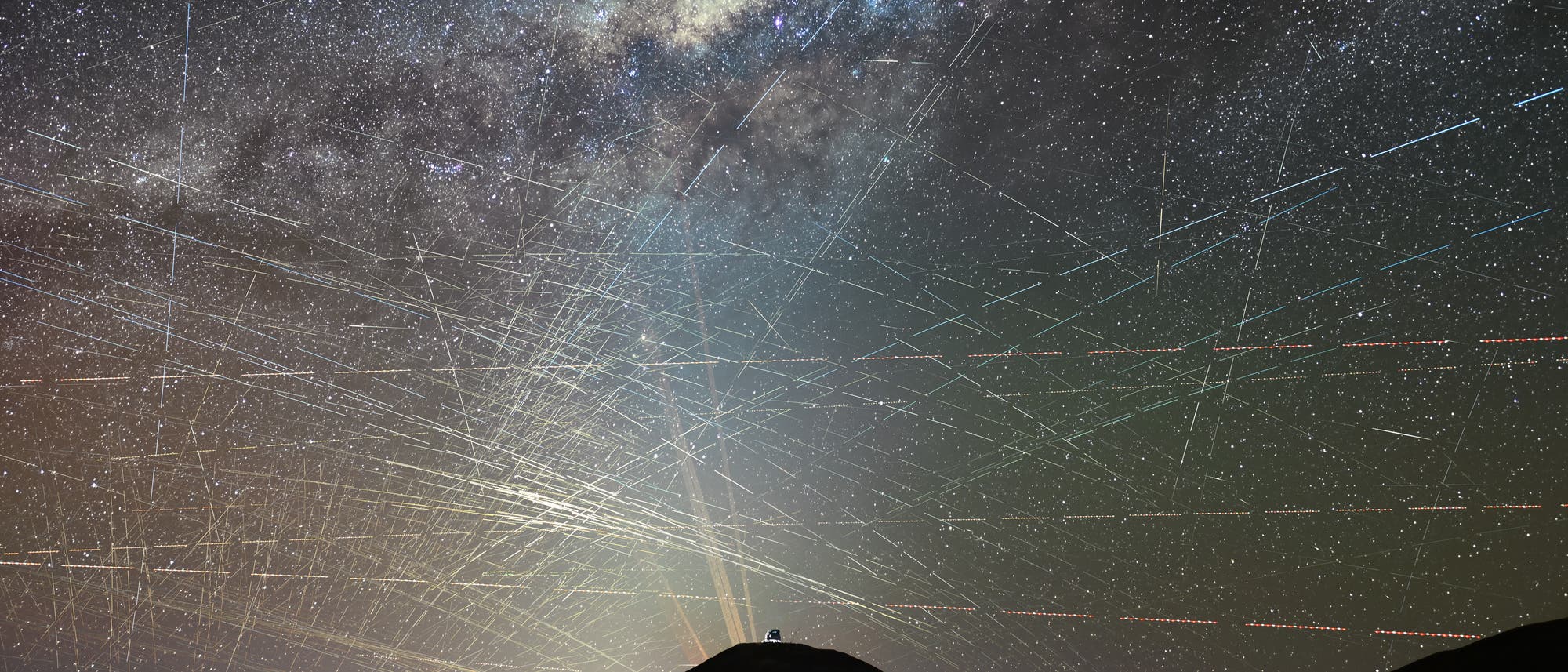 Eine nächtliche Himmelsaufnahme zeigt die Milchstraße, die sich über den Himmel erstreckt. Zahlreiche helle Linien durchziehen das Bild, die Satellitenbahnen darstellen. Im Vordergrund ist die Silhouette eines Hügels mit einem Observatorium zu sehen. Der Himmel ist mit unzähligen Sternen übersät, die ein beeindruckendes Panorama des Universums bieten. Eine nächtliche Himmelsaufnahme zeigt die Milchstraße, die sich über den Himmel erstreckt. Zahlreiche helle Linien durchziehen das Bild, die Satellitenbahnen darstellen. Im Vordergrund ist die Silhouette eines Hügels mit einem Observatorium zu sehen. Der Himmel ist mit unzähligen Sternen übersät, die ein beeindruckendes Panorama des Universums bieten.