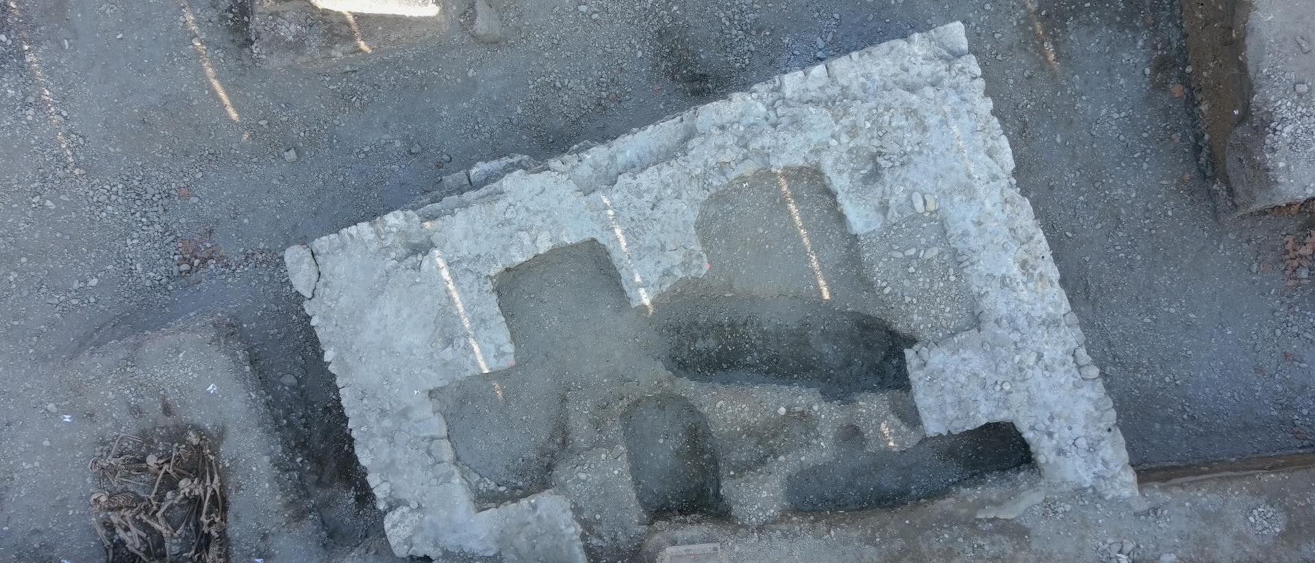 Luftaufnahme einer arch&auml;ologischen Ausgrabungsst&auml;tte mit freigelegten Fundamenten und Strukturen. Die rechteckigen und unregelm&auml;&szlig;igen Formen der Steine deuten auf alte Bauwerke hin. Links im Bild sind &Uuml;berreste von Skeletten zu sehen, die in der Erde eingebettet sind. Der Boden ist &uuml;berwiegend aus grauem Schotter und Erde.