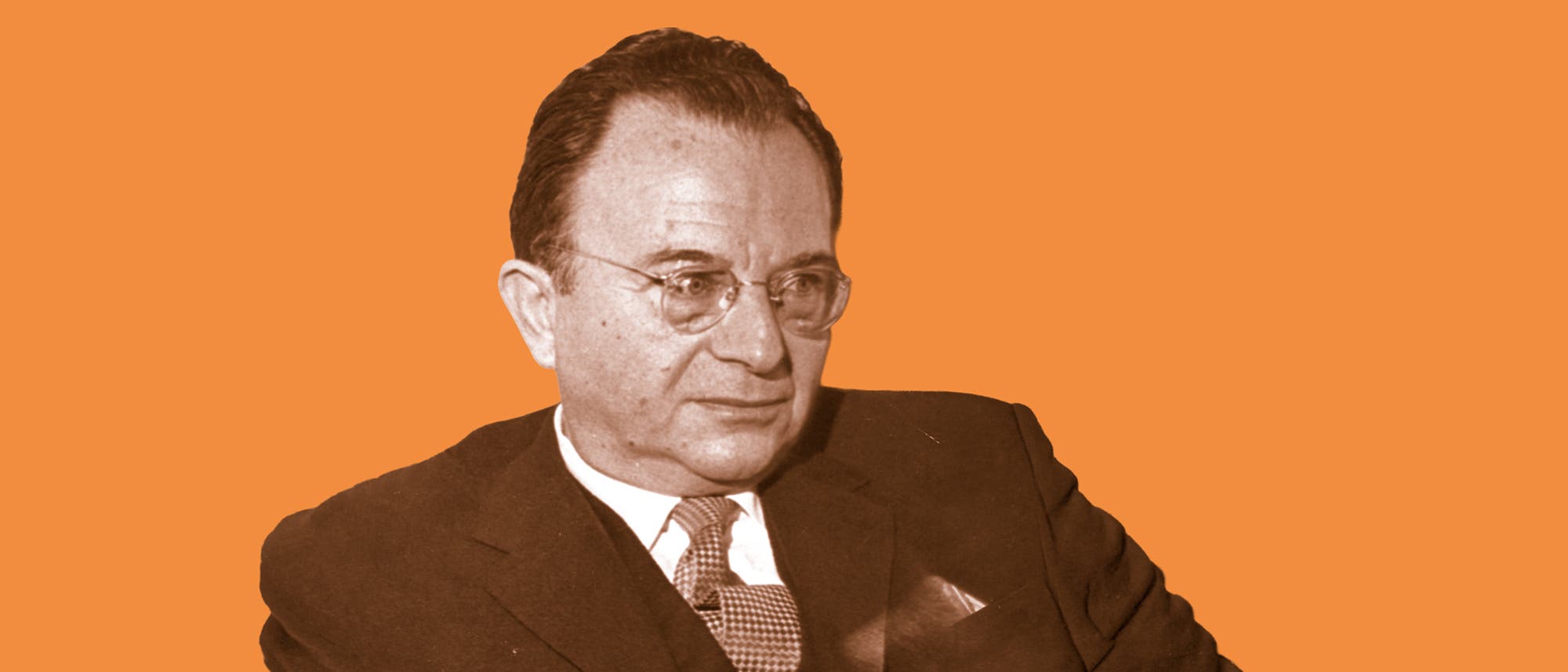 Ein Mann in einem Anzug sitzt vor einem orangefarbenen Hintergrund. Er trägt eine Brille und schaut zur Seite. Das Bild hat einen formellen und professionellen Charakter. Ein Mann in einem Anzug sitzt vor einem orangefarbenen Hintergrund. Er trägt eine Brille und schaut zur Seite. Das Bild hat einen formellen und professionellen Charakter.