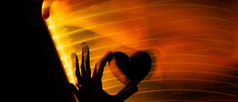 Eine Hand h&auml;lt ein herzf&ouml;rmiges Objekt vor einem Hintergrund aus leuchtenden, wirbelnden Lichtstreifen in warmen Gelb- und Oranget&ouml;nen. Die Szene erzeugt einen kontrastreichen Effekt zwischen der dunklen Silhouette der Hand und den hellen, dynamischen Lichtmustern.