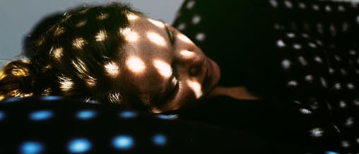 Eine Person liegt entspannt auf einem Kissen, während Sonnenlicht durch ein Fenster mit einem gemusterten Vorhang fällt und ein Muster aus Lichtpunkten auf ihr Gesicht und den Hintergrund wirft. Die Szene vermittelt eine ruhige und friedliche Atmosphäre. Eine Person liegt entspannt auf einem Kissen, während Sonnenlicht durch ein Fenster mit einem gemusterten Vorhang fällt und ein Muster aus Lichtpunkten auf ihr Gesicht und den Hintergrund wirft. Die Szene vermittelt eine ruhige und friedliche Atmosphäre.