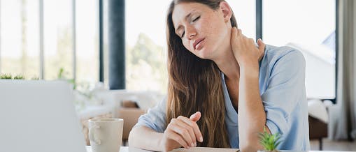 Eine Frau sitzt an einem Schreibtisch mit einem Laptop, hält ihren Nacken und sieht erschöpft aus. Auf dem Tisch liegen ein Notizbuch, ein Stift, eine Tasse und eine kleine Pflanze. Im Hintergrund sind große Fenster mit Blick auf eine verschwommene Landschaft zu sehen. Die Szene vermittelt das Gefühl von Müdigkeit oder Nackenschmerzen während der Arbeit.
