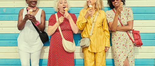 Vier gut gelaunte Frauen mittleren Alters lehnen an einer blau-wei&szlig; gestreiften Wand und essen Eis. Die Frau links tr&auml;gt ein wei&szlig;es Kleid, die Frau daneben ein gepunktetes rotes Kleid, die dritte einen gelben Jumpsuit und die vierte ein gebl&uuml;mtes Kleid. Alle lachen und scheinen das Leben zu genie&szlig;en.