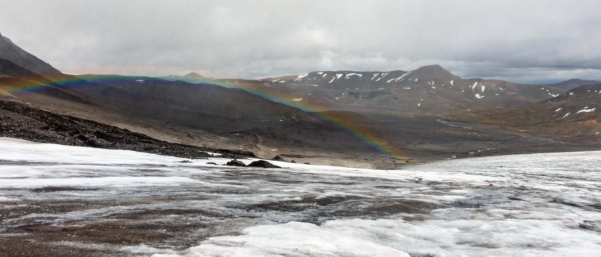Eine Gletscherlandschaft mit schneebedecktem Vordergrund und einem Regenbogen, der sich über das Tal erstreckt. Im Hintergrund sind schroffe Berggipfel unter bewölktem Himmel zu sehen. Die Szene vermittelt eine ruhige, natürliche Schönheit. Eine Gletscherlandschaft mit schneebedecktem Vordergrund und einem Regenbogen, der sich über das Tal erstreckt. Im Hintergrund sind schroffe Berggipfel unter bewölktem Himmel zu sehen. Die Szene vermittelt eine ruhige, natürliche Schönheit.