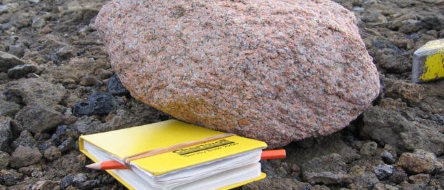 Ein großer, rötlich-brauner Felsbrocken liegt auf einer felsigen Oberfläche. Vor dem Felsbrocken befindet sich ein gelbes Notizbuch mit einem roten Stift, das auf dem Boden liegt. Im Hintergrund ist ein Gewässer zu sehen. Die Szene vermittelt einen Eindruck von Feldforschung oder geologischen Studien. Ein großer, rötlich-brauner Felsbrocken liegt auf einer felsigen Oberfläche. Vor dem Felsbrocken befindet sich ein gelbes Notizbuch mit einem roten Stift, das auf dem Boden liegt. Im Hintergrund ist ein Gewässer zu sehen. Die Szene vermittelt einen Eindruck von Feldforschung oder geologischen Studien.