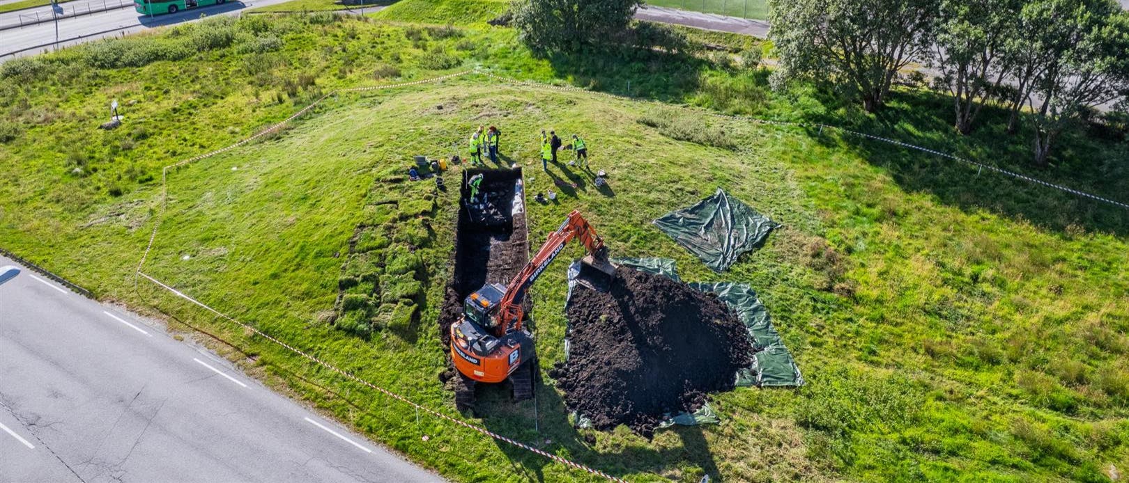 Luftaufnahme einer arch&auml;ologischen Ausgrabungsst&auml;tte auf einer gr&uuml;nen Wiese neben einer Stra&szlig;e. Eine orangefarbene Baggermaschine hebt Erde aus einem rechteckigen Graben. Mehrere Personen in Sicherheitskleidung arbeiten an der Ausgrabungsstelle. Im Hintergrund sind ein Fluss, Geb&auml;ude und ein vorbeifahrender Bus zu sehen.