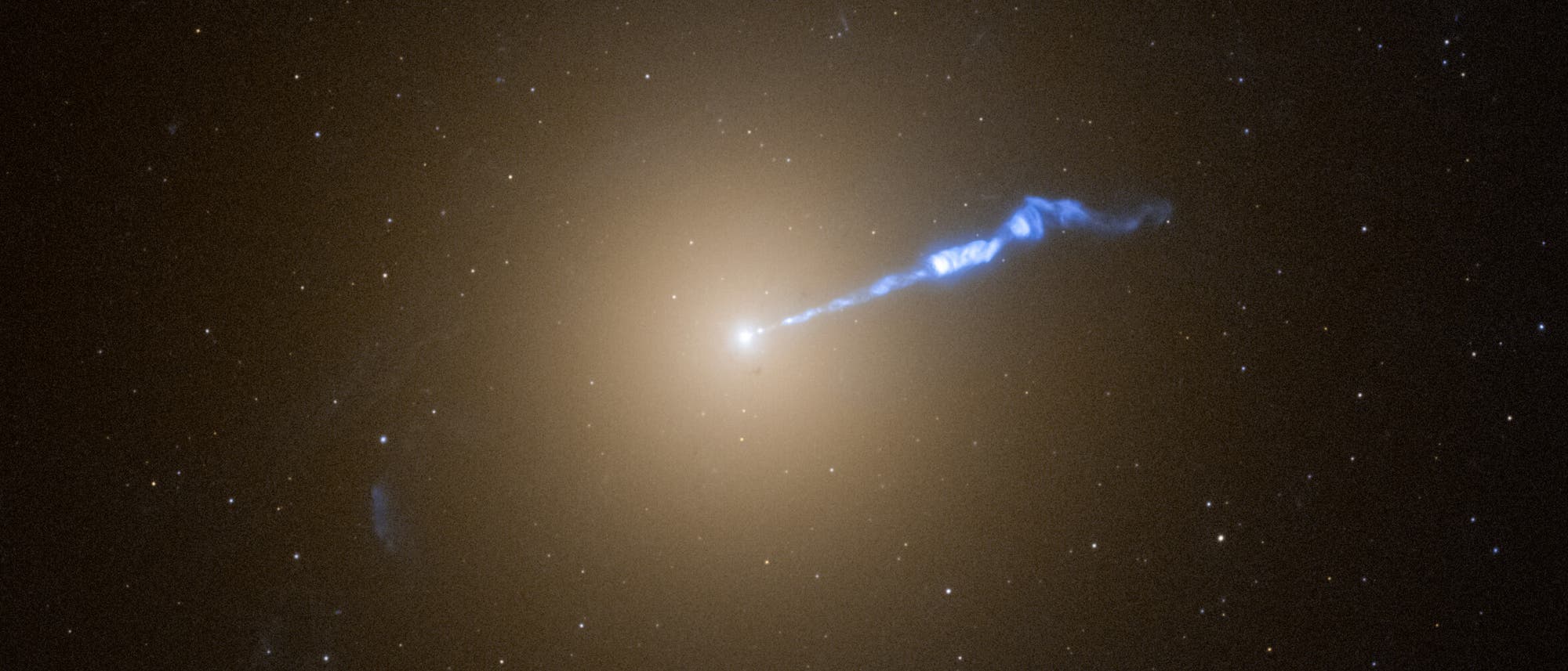 Ein astronomisches Bild zeigt eine Galaxie mit einem hellen, zentralen Kern, aus dem ein blauer Plasmastrahl hervorgeht. Der Strahl erstreckt sich nach rechts und hebt sich vor dem dunklen Hintergrund des Weltraums ab, der mit zahlreichen kleinen Sternen &uuml;bers&auml;t ist. Der zentrale Bereich der Galaxie erscheint orangefarben und diffus. Das Bild veranschaulicht die Dynamik und Struktur von Galaxienjets im Universum.