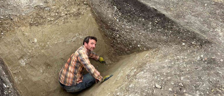 Eine Person in einem karierten Hemd und Handschuhen gräbt in einer archäologischen Ausgrabungsstätte. Sie befindet sich in einer tiefen, spitzförmigen Grube und verwendet ein kleines Werkzeug, um den Boden zu bearbeiten. Die Umgebung zeigt mehrere Erdschichten und freigelegte Bereiche, die auf eine systematische Untersuchung hindeuten. Eine Person in einem karierten Hemd und Handschuhen gräbt in einer archäologischen Ausgrabungsstätte. Sie befindet sich in einer tiefen, spitzförmigen Grube und verwendet ein kleines Werkzeug, um den Boden zu bearbeiten. Die Umgebung zeigt mehrere Erdschichten und freigelegte Bereiche, die auf eine systematische Untersuchung hindeuten.