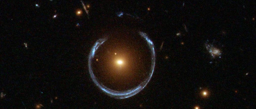 Ein Bild eines Einsteinrings, einer astronomischen Erscheinung, bei der das Licht einer entfernten Galaxie durch die Gravitationskraft einer näheren Galaxie gebogen wird, wodurch ein ringförmiges Muster entsteht. Im Zentrum des Bildes befindet sich eine helle, gelbliche Galaxie, umgeben von einem bläulichen Lichtbogen. Mehrere kleinere Lichtpunkte, die entfernte Sterne oder Galaxien darstellen, sind im Hintergrund zu sehen. Das Bild veranschaulicht das Phänomen der Gravitationslinsen in der Astronomie.