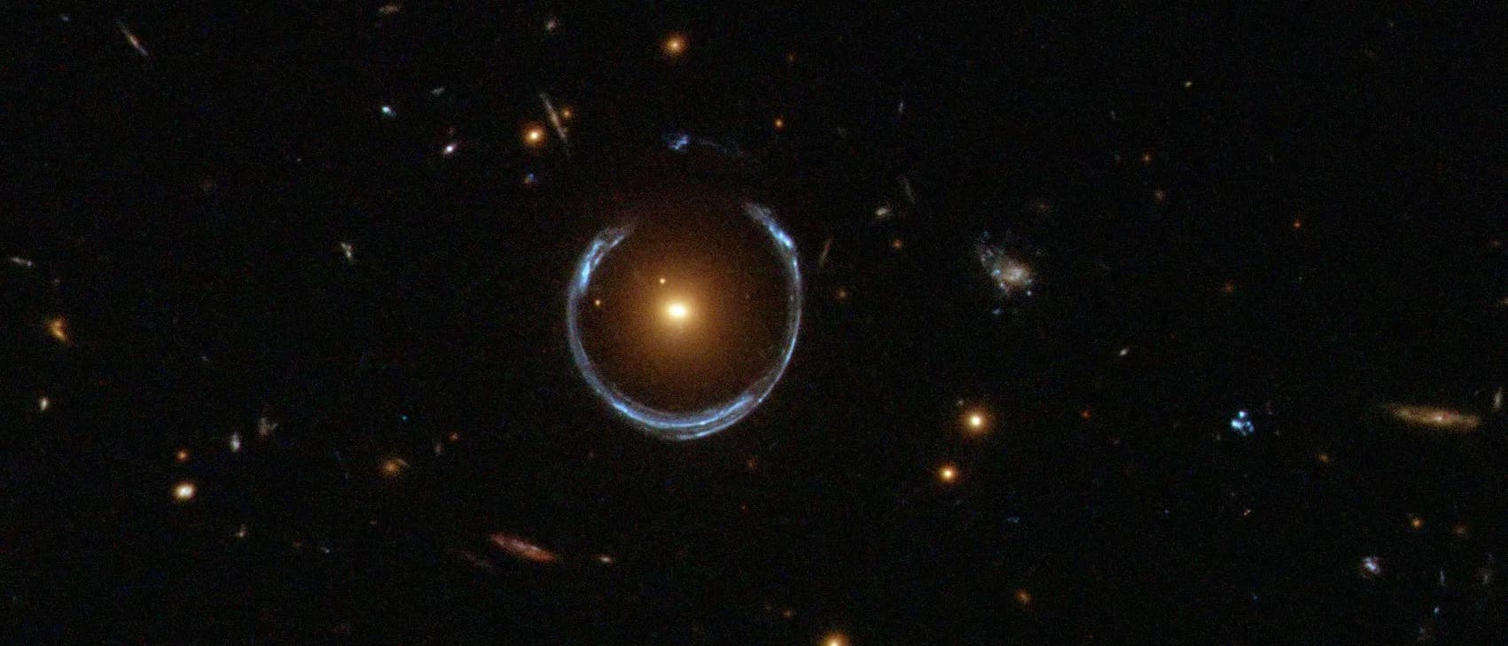 Ein astronomisches Bild zeigt ein Ph&auml;nomen namens Einsteinring. Im Zentrum befindet sich eine helle, orangefarbene Galaxie, umgeben von einem verzerrten, bl&auml;ulichen Lichtbogen, der durch Gravitationslinseneffekte entsteht. Mehrere kleinere, leuchtende Punkte und Galaxien sind im Hintergrund sichtbar. Das Bild veranschaulicht die Wirkung der Schwerkraft auf das Licht entfernter Objekte im Universum.