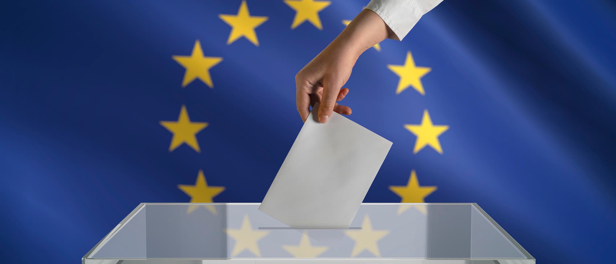 Eine Hand wirft einen Zettel in einen durchsichtigen Kasten. Im Hintergrund die Europaflagge.