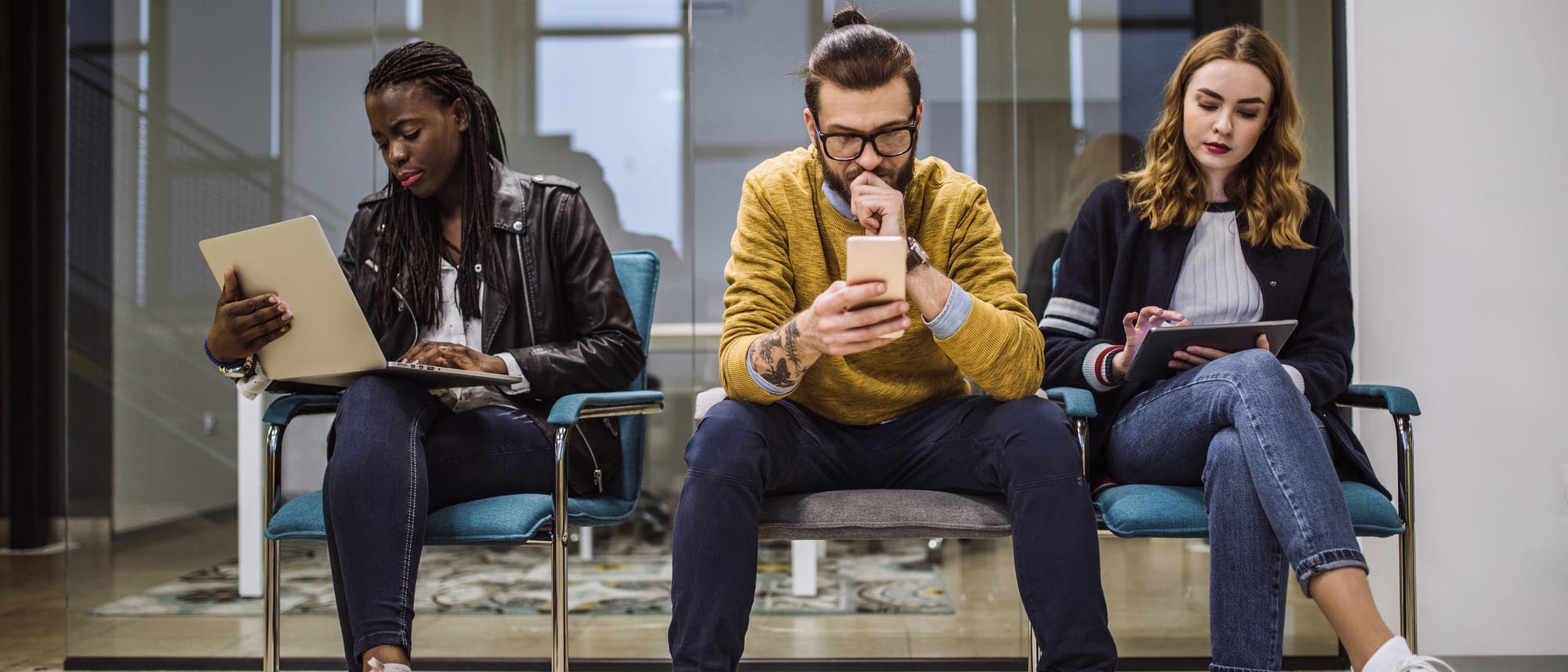 Drei Studierende sitzen auf Stühlen und beschäftigen sich mit Handy, Tablet oder Computer