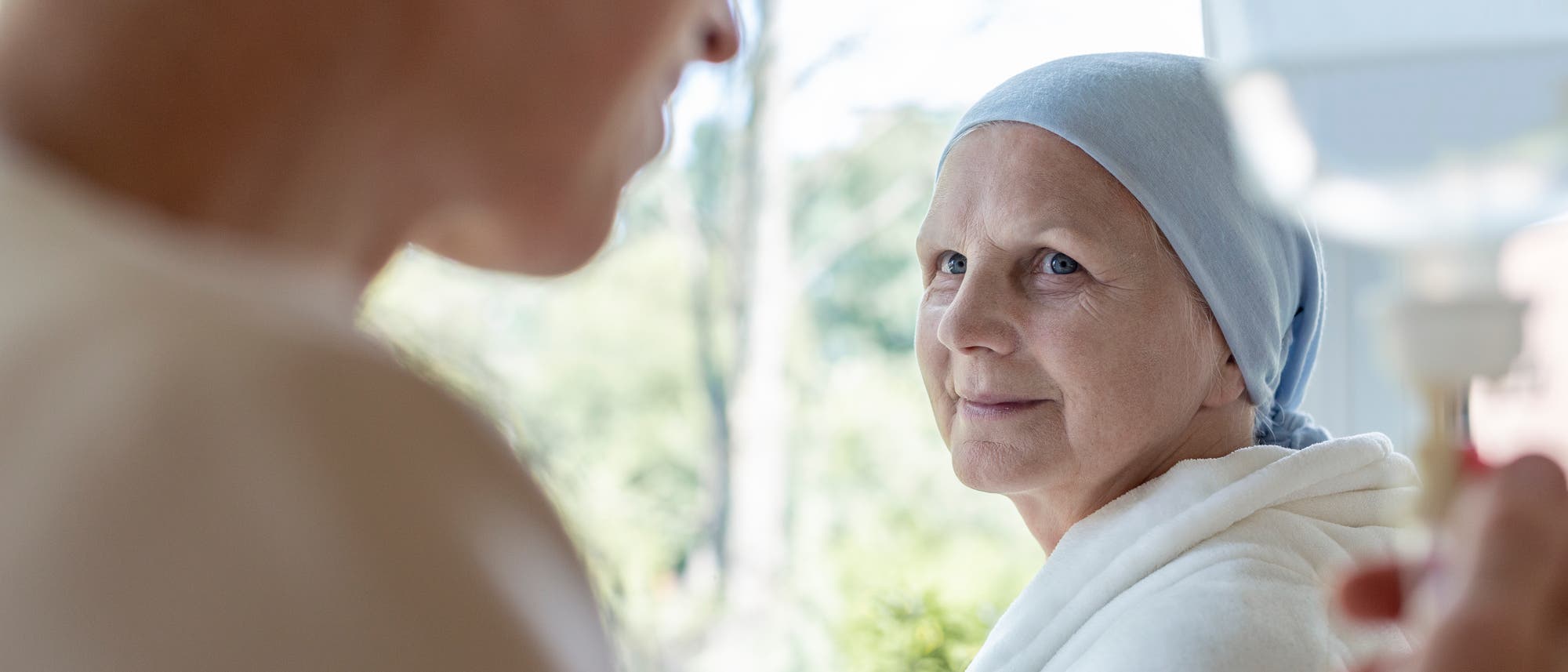 Eine offenbar an Krebs erkrankte Frau trägt ein Kopftuch und schaut hoffnungsvoll eine andere Frau an, die gerade einen Infusionsbeutel reguliert.