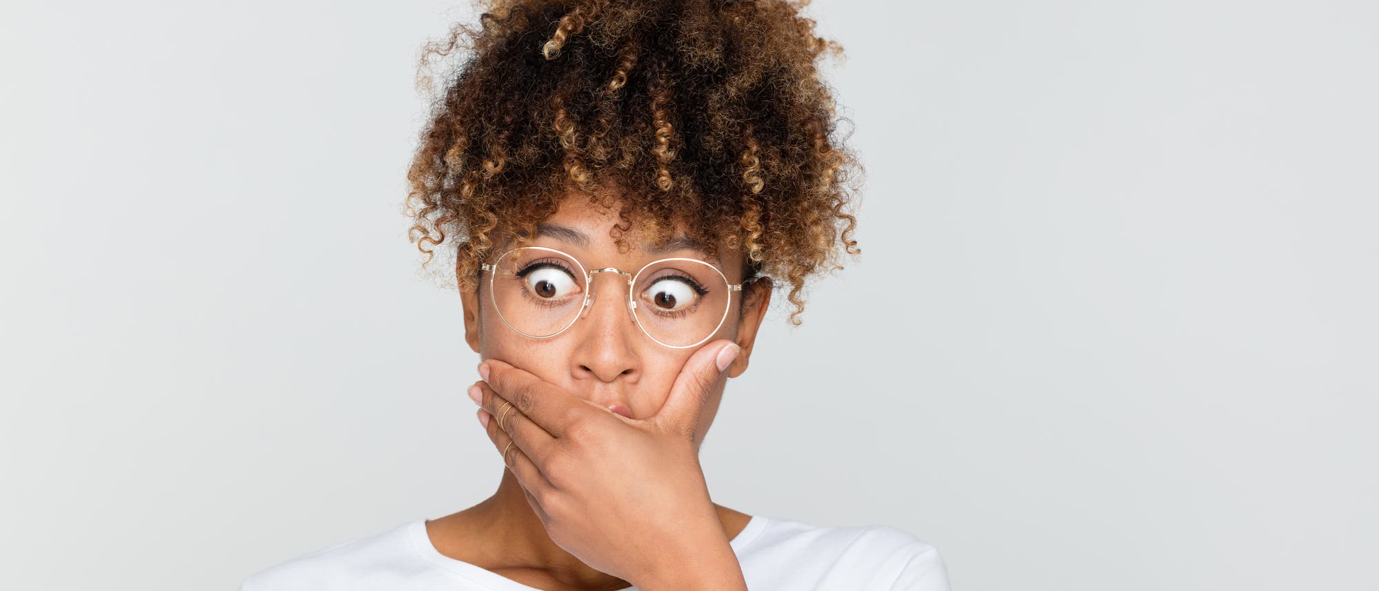 Eine Person mit lockigem Haar und Brille h&auml;lt sich &uuml;berrascht die Hand vor den Mund. Sie tr&auml;gt ein wei&szlig;es Oberteil und blickt mit weit ge&ouml;ffneten Augen in die Kamera. Der Hintergrund ist einfarbig hellgrau.