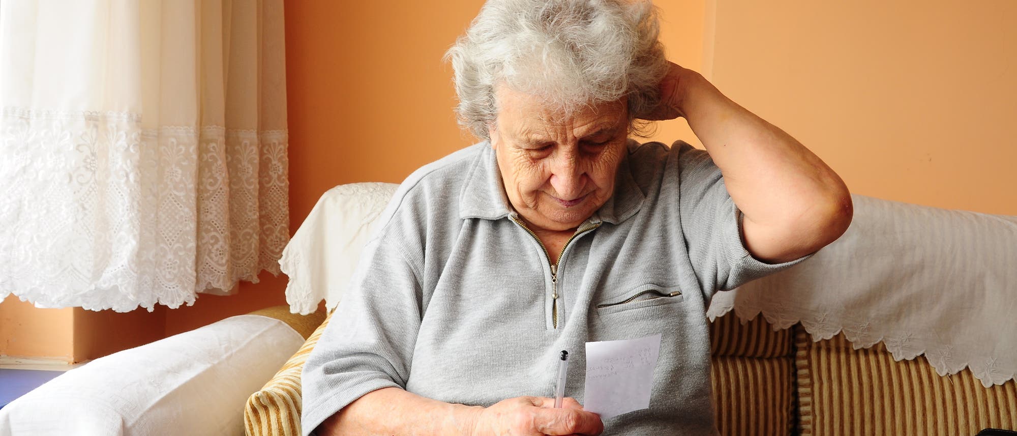 Eine ältere Dame schaut auf einen Zettel und scheint sich den Kopf zu reiben.