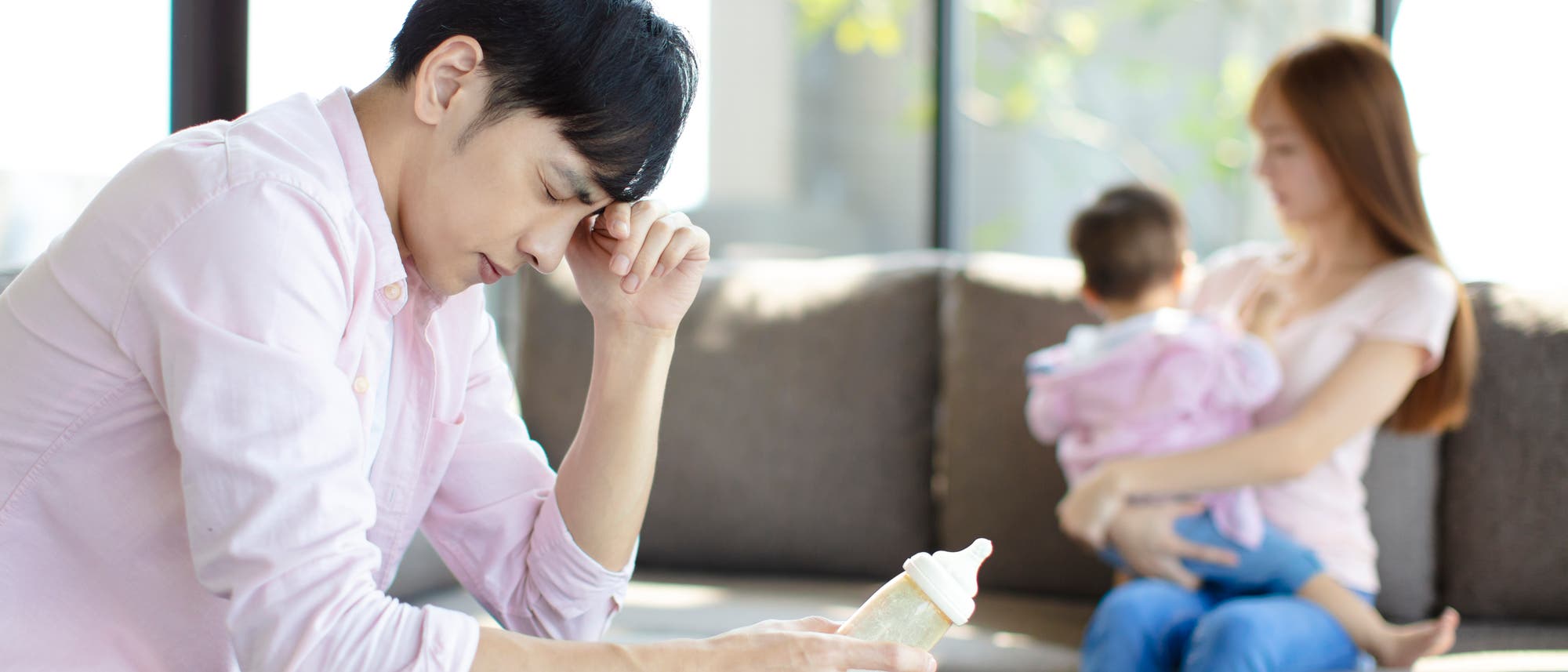 Ein erschöpft wirkender Vater sitzt auf dem Sofa, im Hintergrund sind die Mutter mit einem Baby zu sehen