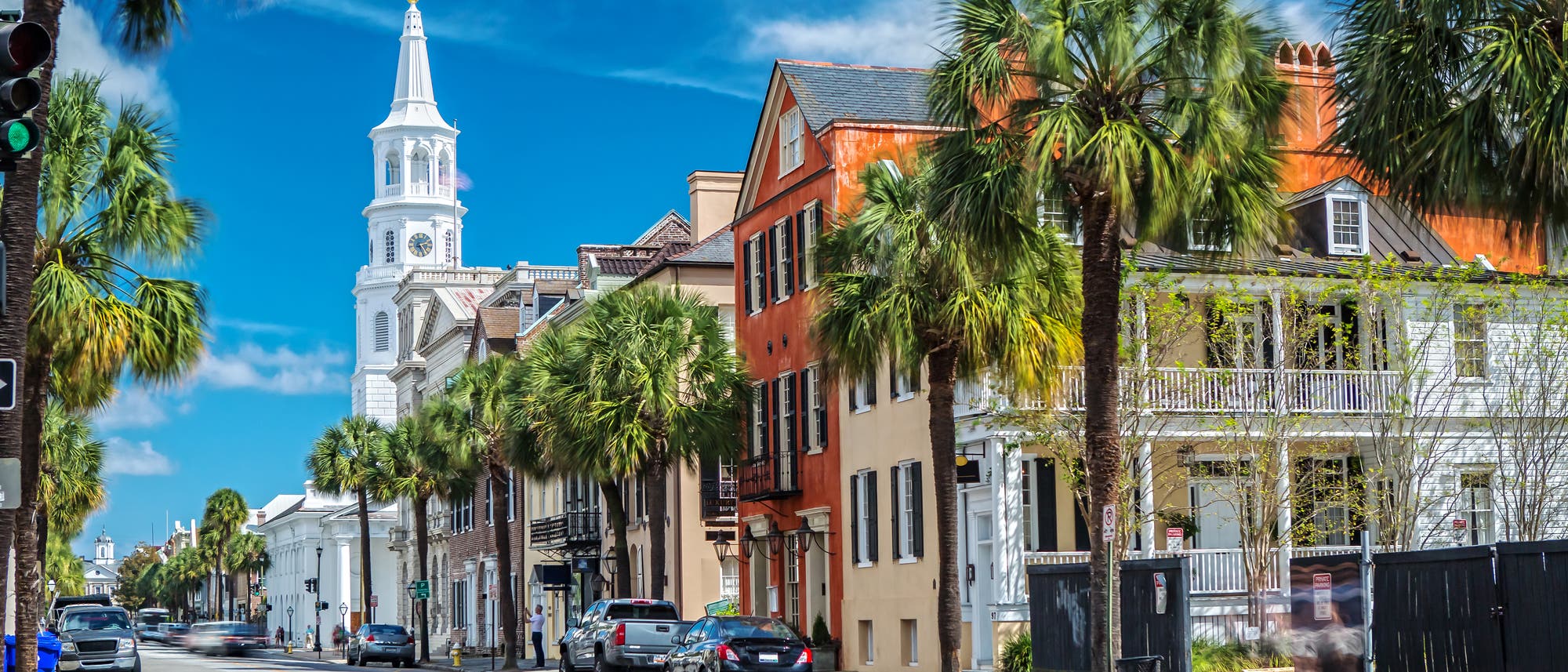 Straßenszene aus Charleston, South Carolina: Im Hintergrund die weiße St.-Michaels-Kirche, der Himmel ist blau, Palmen säumen den Straßenrand.