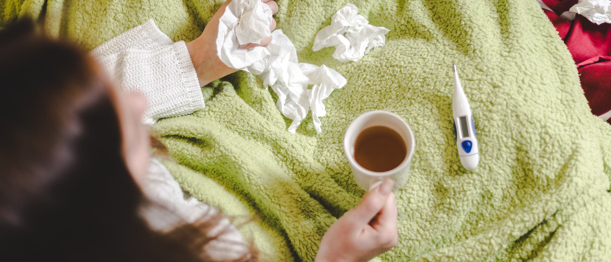 Eine Frau mit den üblichen Erkältungsutensilien: Decke, Tee, Taschentücher, Fieberthermometer.