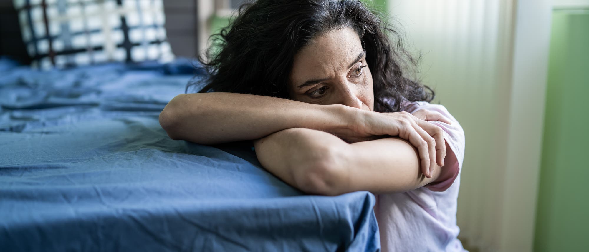Eine Person sitzt nachdenklich auf dem Boden neben einem Bett mit blauer Bettwäsche. Sie stützt ihren Kopf auf die Arme, die auf der Bettkante ruhen. Im Hintergrund ist ein Kissen mit Karomuster zu sehen. Der Raum hat grüne Wände und ein Fenster, durch das Licht einfällt.