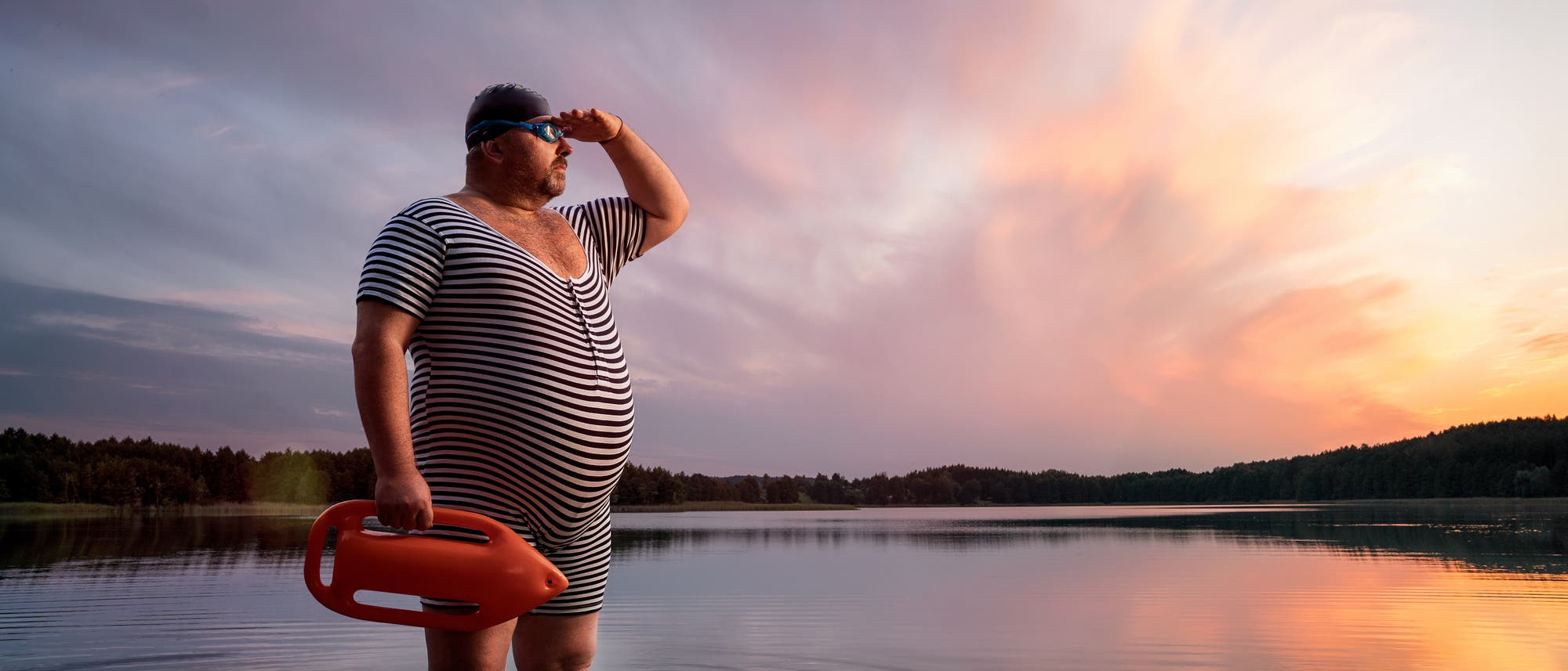 Ein etwas älterer, wohlbeleibter Rettungsschwimmer in gestreiftem Badeanzug steht bei Sonnenuntergang im Wasser eines Sees.