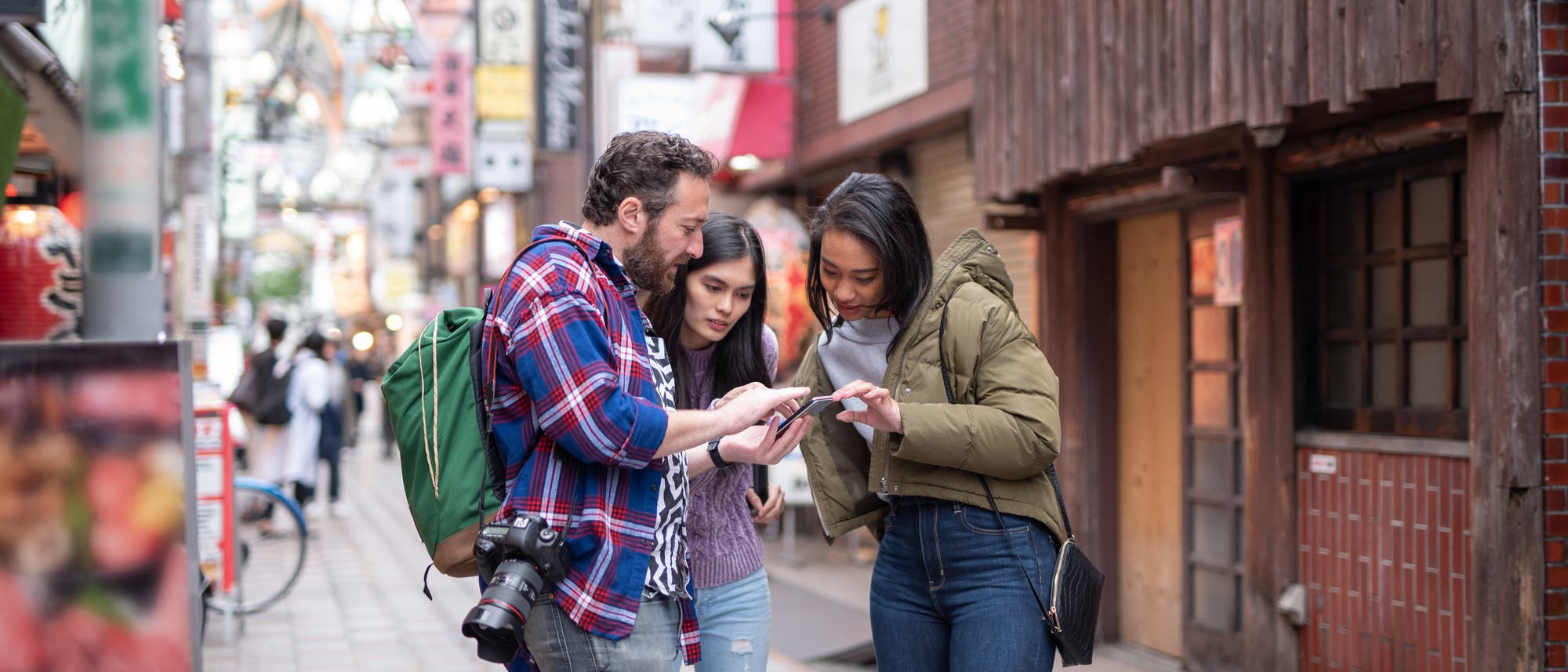 Zwei Touristen fragen eine Einheimische in den Straßen von Tokio, dessen bunte Schilder man im Hintergrund sieht, mit dem Smartphone nach dem Weg.