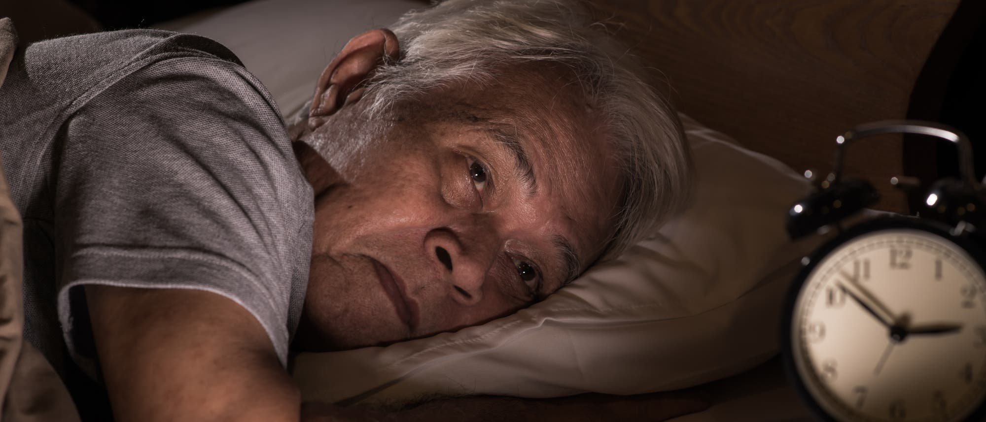 Älterer Mann liegt schlaflos im Bett und der Wecker zeigt drei Uhr morgens.