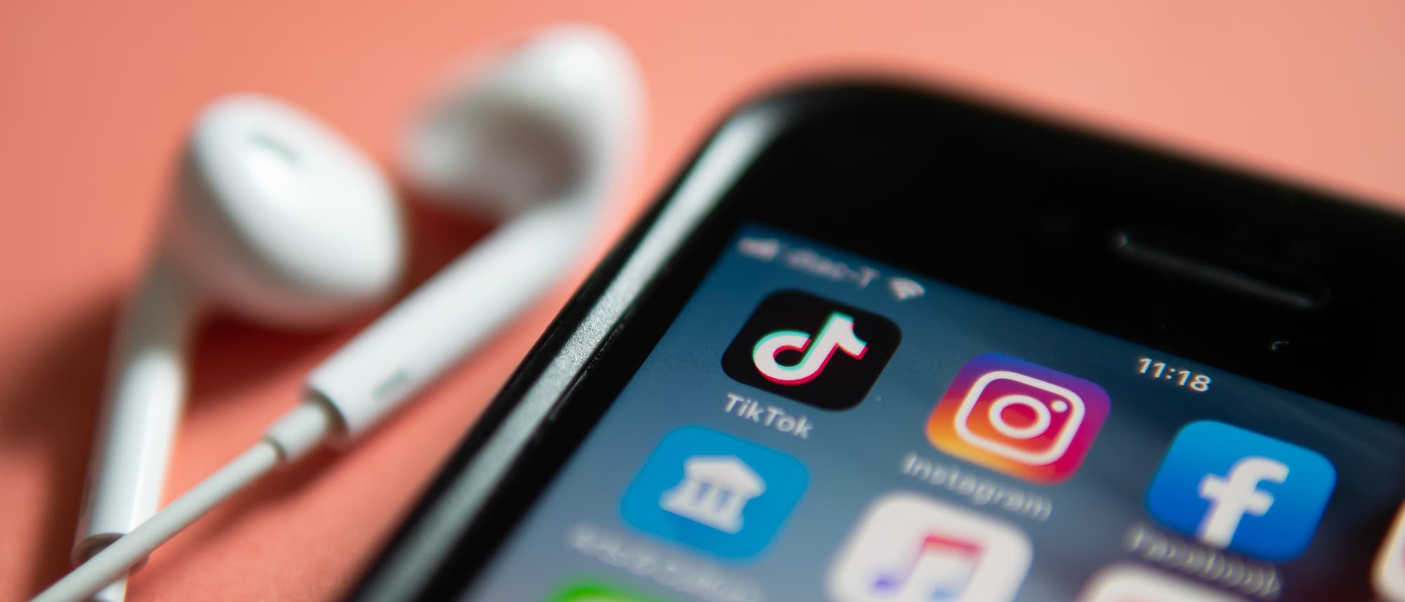 Smartphone in der Nahaufnahme vor rosafarbenem Hintergrund. Auf dem Bildschirm sind die Symbole verschiedener Apps zu sehen, darunter die von TikTok und Instagram. Neben dem Smartphone befinden sich weiße In-Ear-Kopfhörer.
