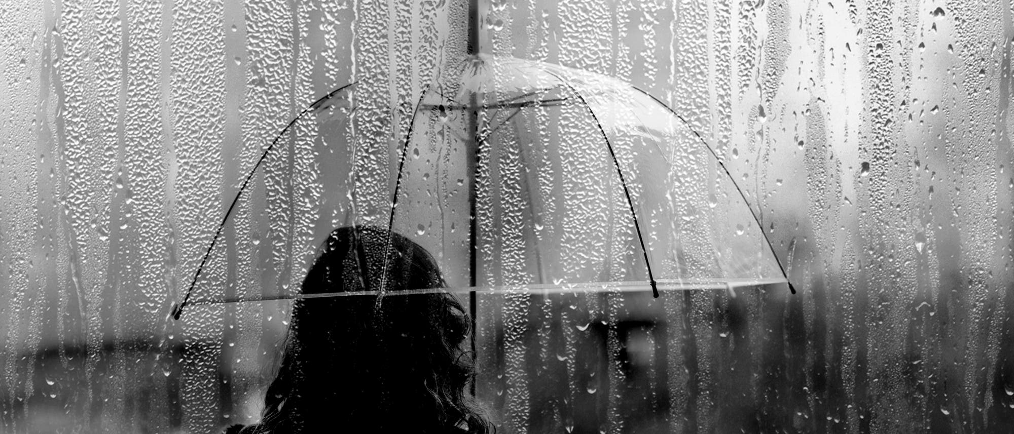 Schwarz-Weiß-Bild einer Frau, die mit einem durchsichtigen Regenschirm im Regen steht. Man sieht sie nur von hinten. 