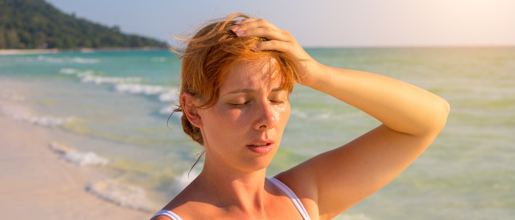 Frau steht an einem Strand in der Sonne, sieht erschöpft aus und fasst sich an den Kopf. 