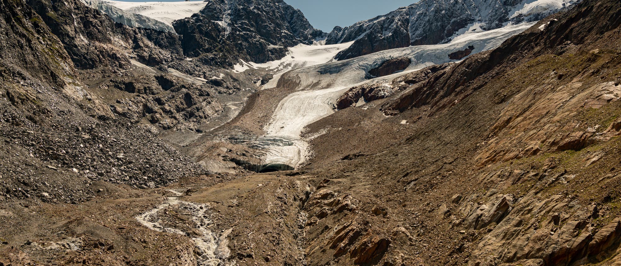 Eine Schotterfläche mit einer kleinen Eiszunge und Bergen im Hintergrund.