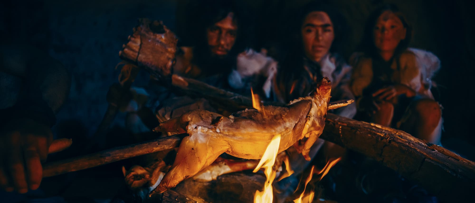 Drei als Frühmenschen verkleidete Homo sapiens braten Tierfleisch über dem Feuer.