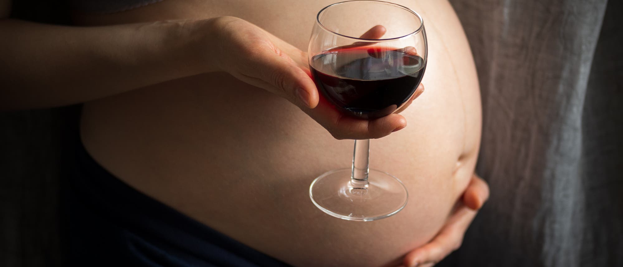 Die Körpermitte einer Schwangeren, von der Seite fotografiert, mit einem Weinglas in der Hand mittig im Bild.