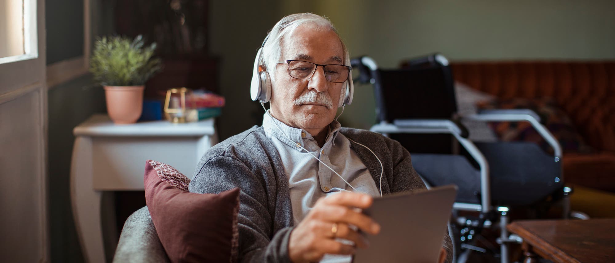 Älterer Mann mit Kopfhörern sitzt in einem Sessel und benutzt ein Tablet.