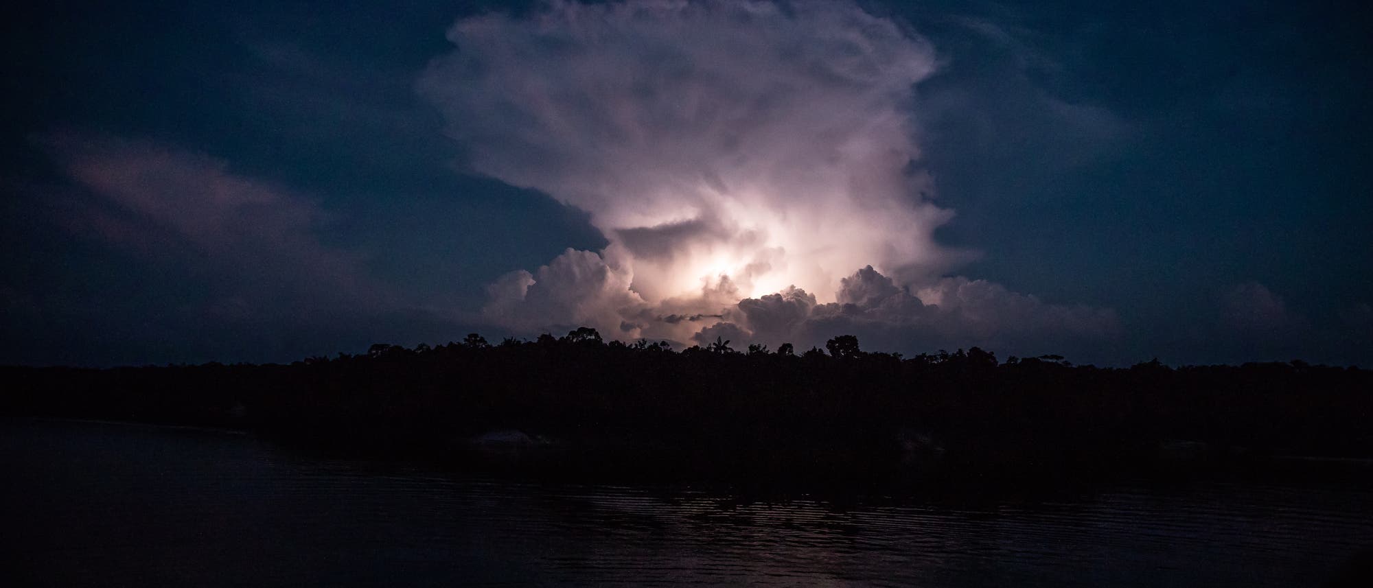 Ein Blitz erleuchtet eine Gewitterwolke in der Nacht über dem Amazonasregenwald, Bäume sind als dunkle Silhouetten zu sehen