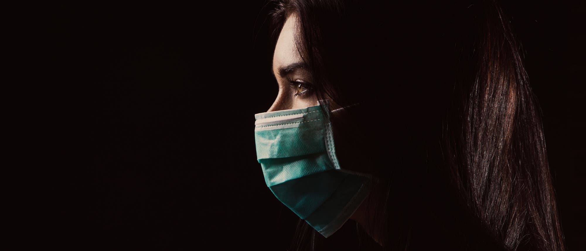 Frau mit medizinischer Mund-Nase-Maske im Profil vor dunklem Hintergrund.
