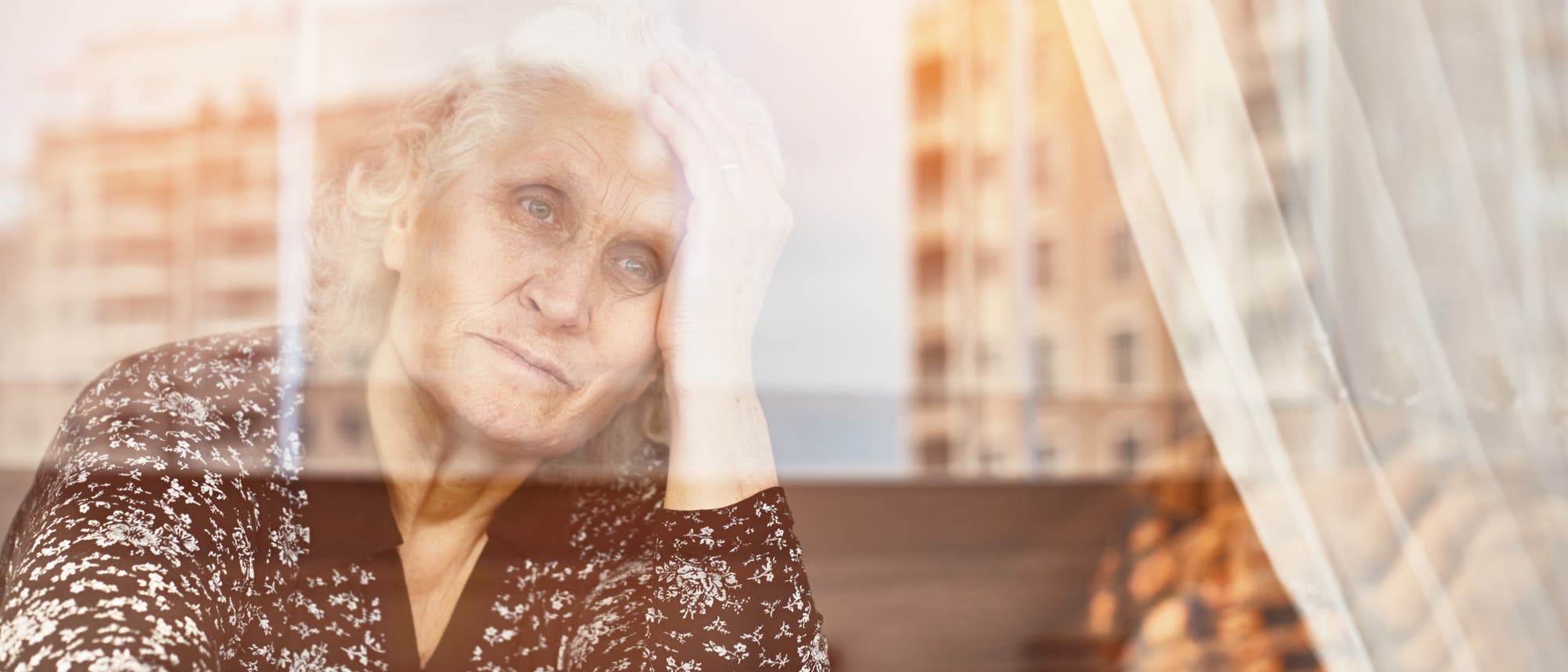 Eine &auml;ltere Frau schaut traurig aus ihrem Fenster. Die Farben sind blass. In der Fensterspiegelung erkennt man triste Hochh&auml;user.