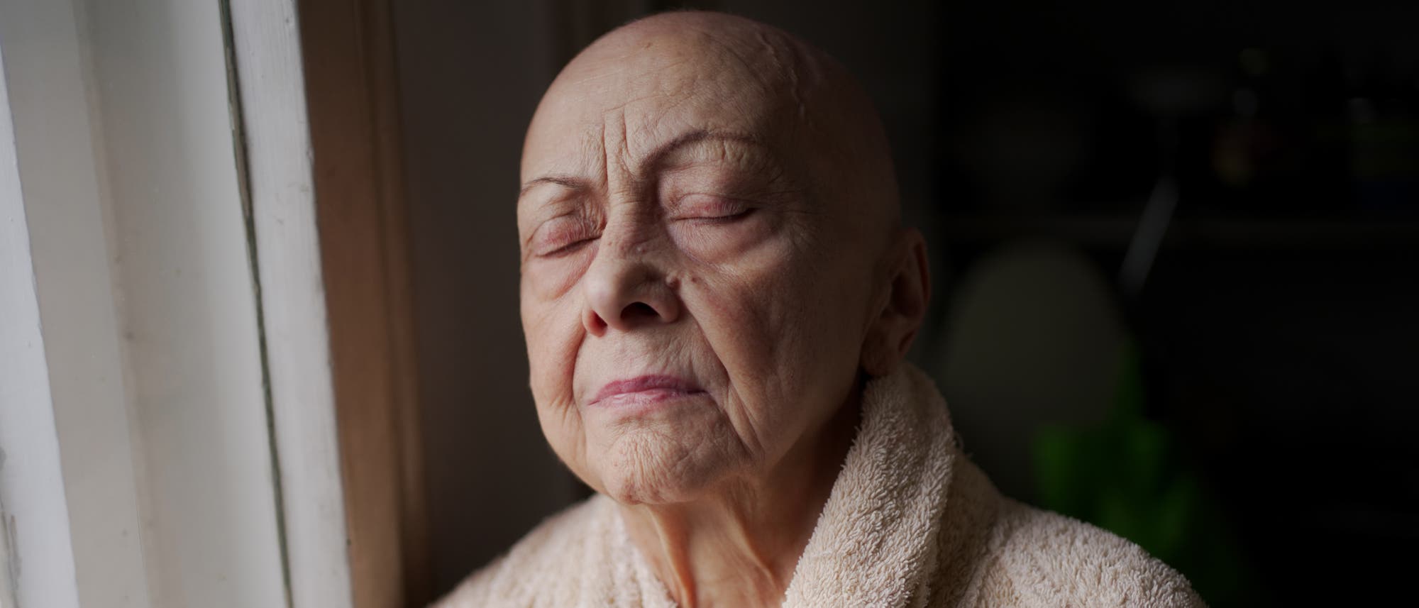 Porträt einer älteren Frau ohne Haare, die im Bademantel am Fenster steht und dabei die Augen geschlossen hat