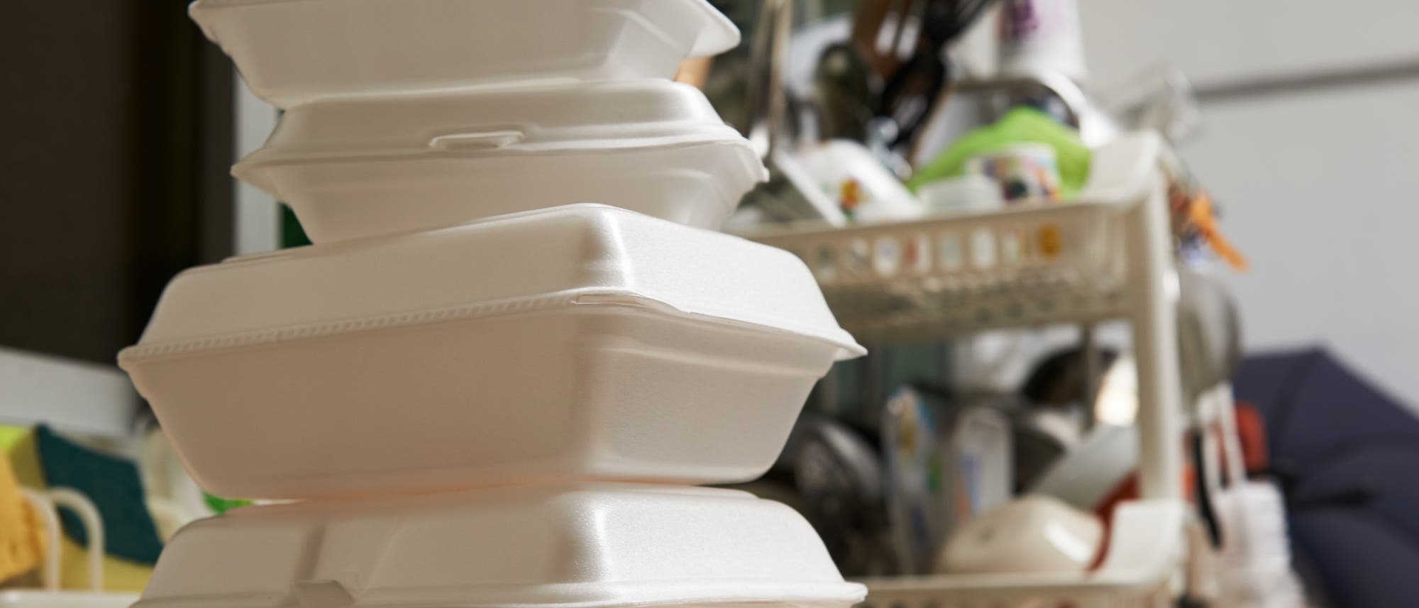 Styroporverpackungen von gelieferten Essen in einer Küche, im Hintergrund schmutziges Geschirr.