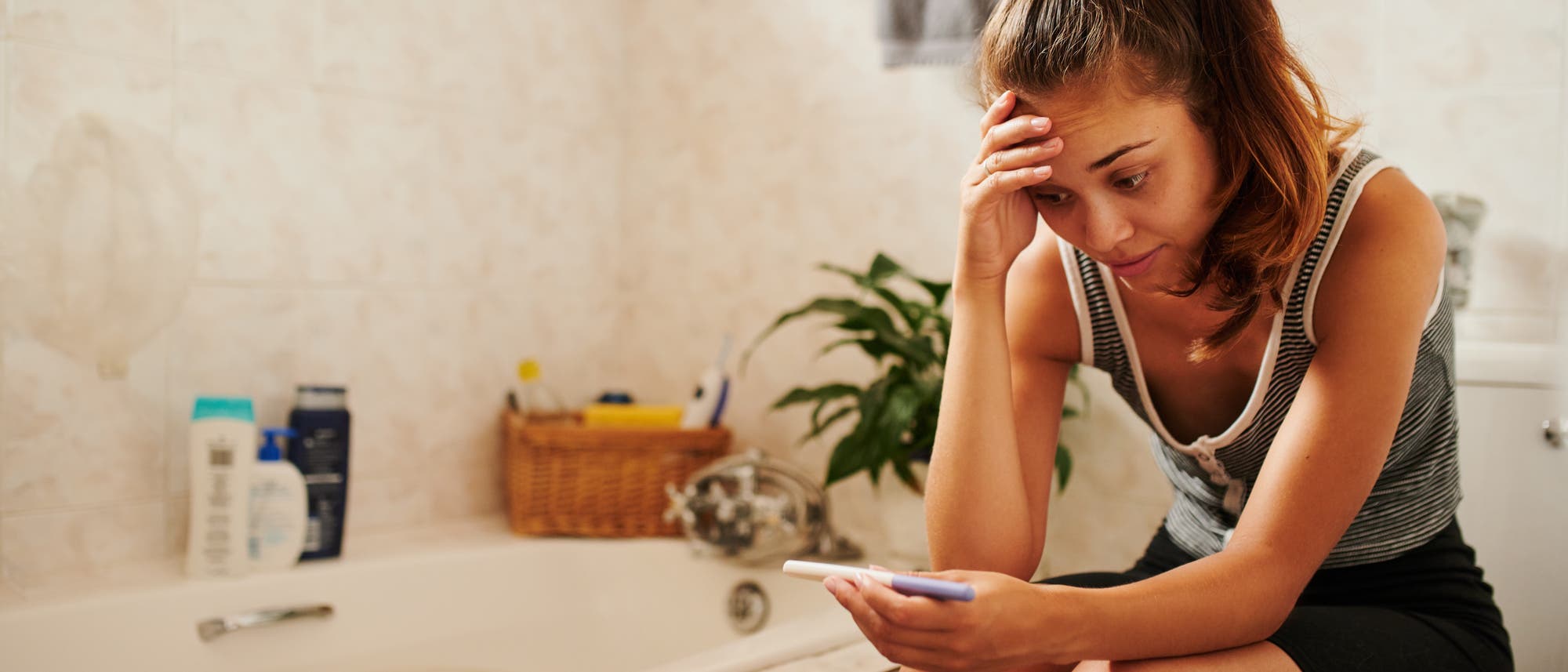 Eine junge Frau sitzt im Bad und schaut ungläubig auf einen Schwangerschaftstest 