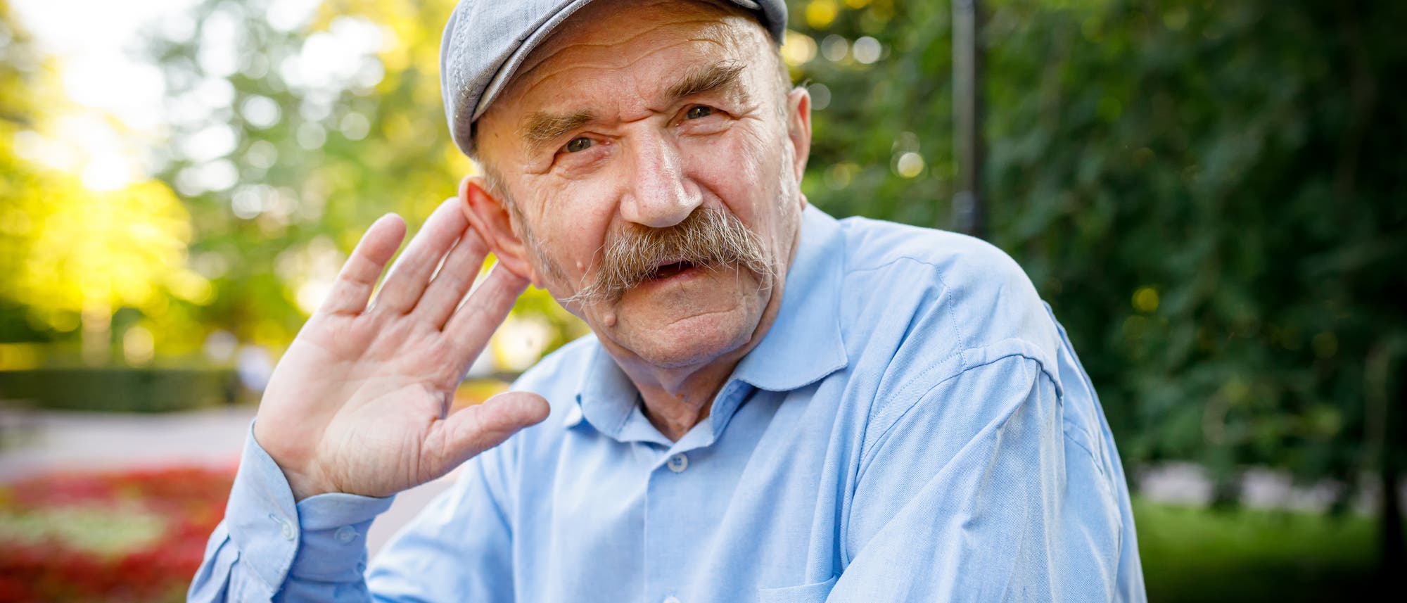 Ein älterer Mann schaut den Betrachter an und hält sich die Hand hinter das Ohr, um besser zu verstehen.