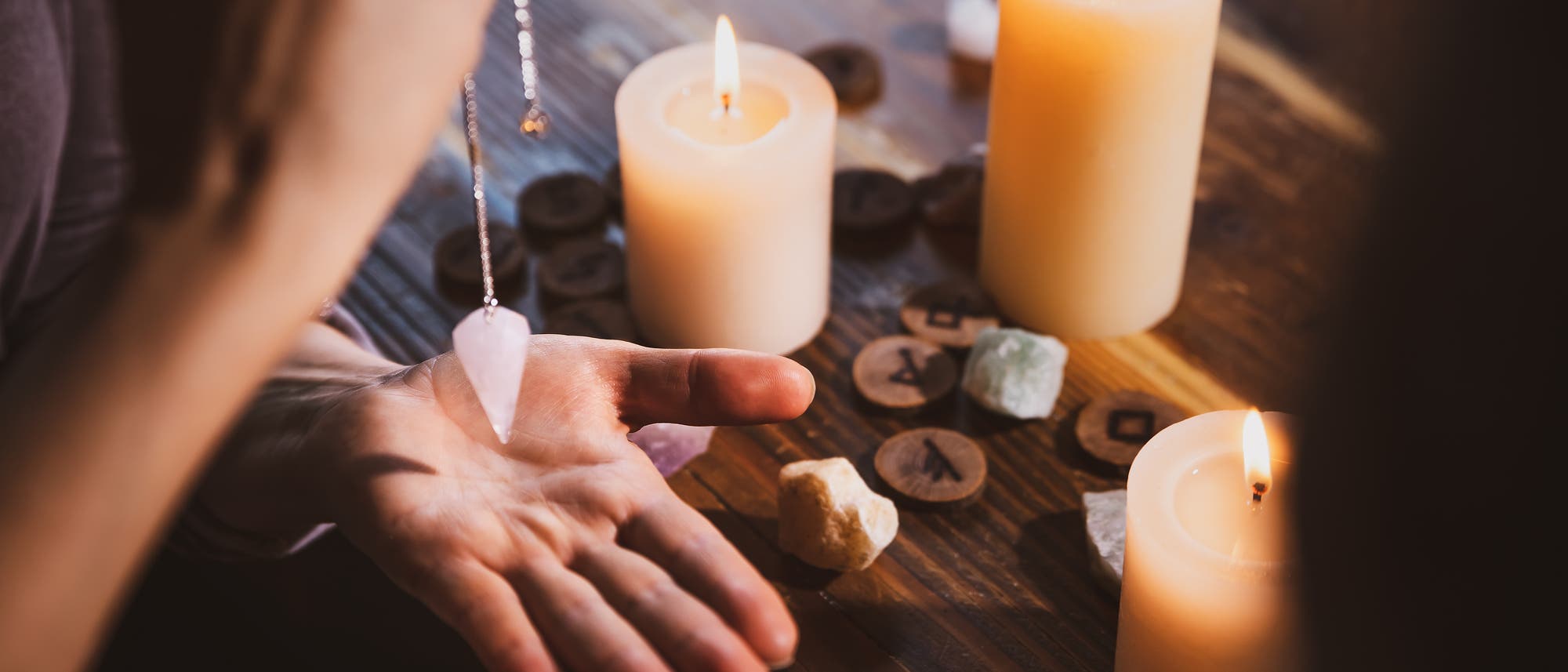 Hand mit Pendel, im Hintergrund stehen Kerzen und bunte Steine liegen auf einem Tisch