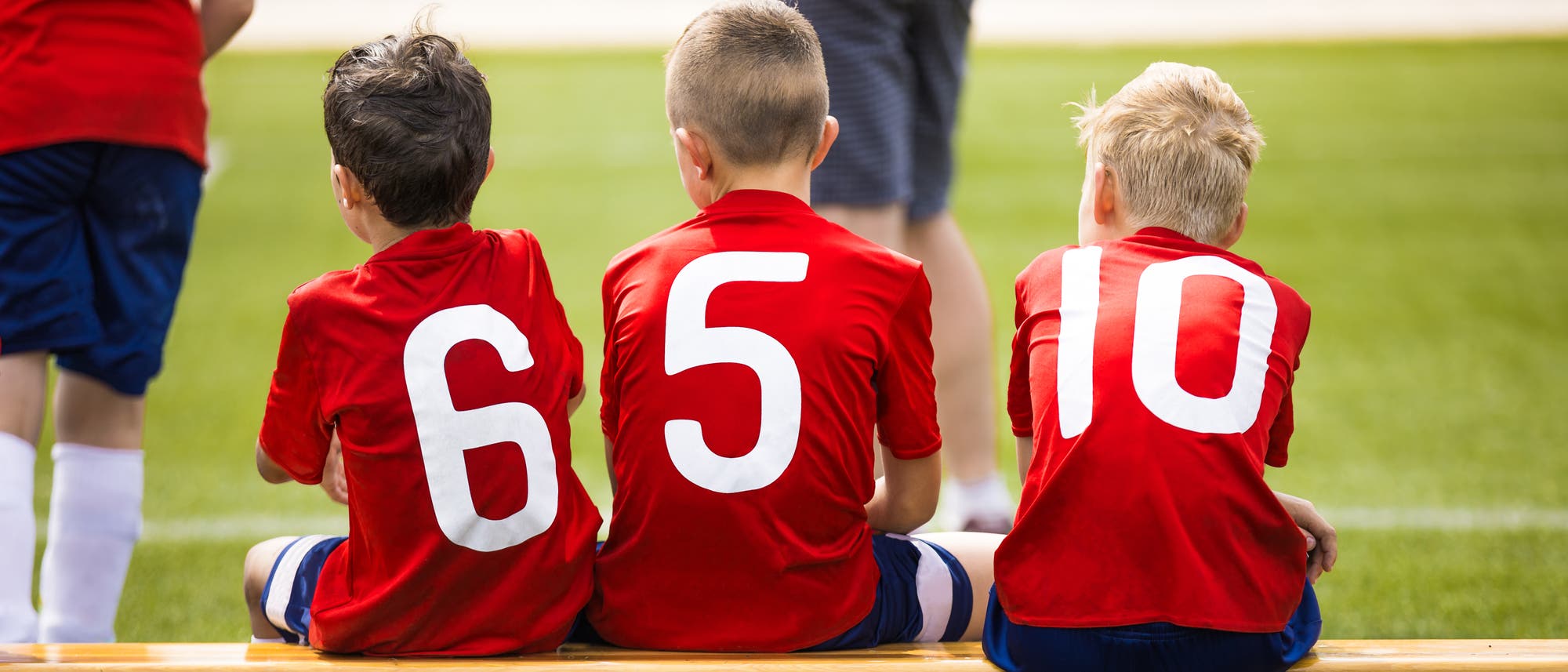 Drei Jungs in Fußballtrikots sitzen auf der Ersatzbank und gucken dem Spiel zu.