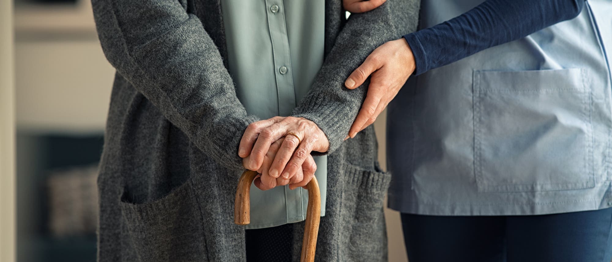 Eine Pflegekraft in blauer Uniform unterstützt eine ältere Person, die einen Gehstock hält, indem sie den Arm der älteren Person stützt. Die Szene vermittelt Fürsorge und Unterstützung im Pflegeumfeld.