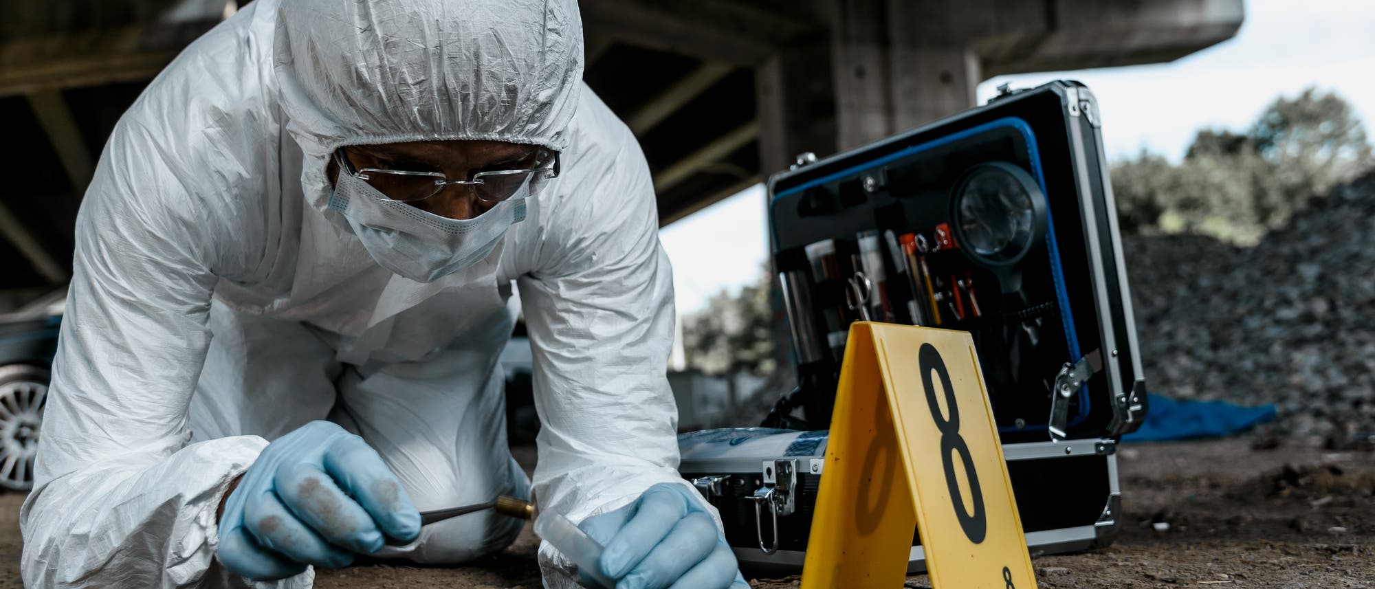 Forensiker im Schutzanzug aus Kunststoff bei der bodennahen Arbeit, vor ihm oder ihr liegen Patronenhülsen. Offenbar fand ein Schusswechsel statt. Der Tatort trägt Nummern. Forensiker im Schutzanzug aus Kunststoff bei der bodennahen Arbeit, vor ihm oder ihr liegen Patronenhülsen. Offenbar fand ein Schusswechsel statt. Der Tatort trägt Nummern.