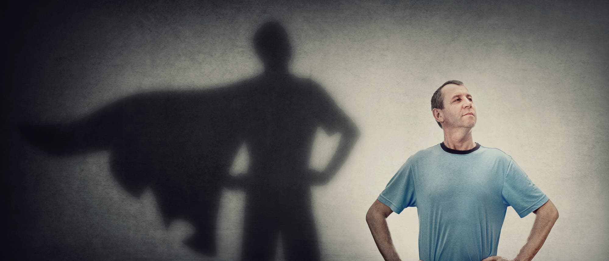 Mann im T-Shirt vor einer Wand; sein Schatten zeigt ihn mit Heldencape.