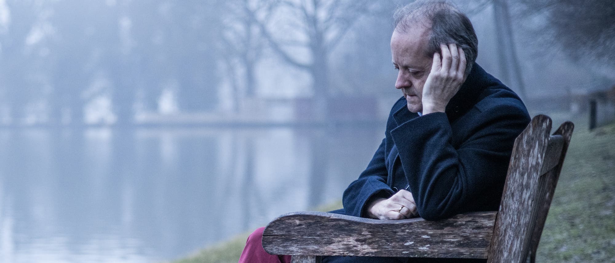 Ein Mann mit grauem Haar sitzt nach nachdenklich auf einer Bank am See 