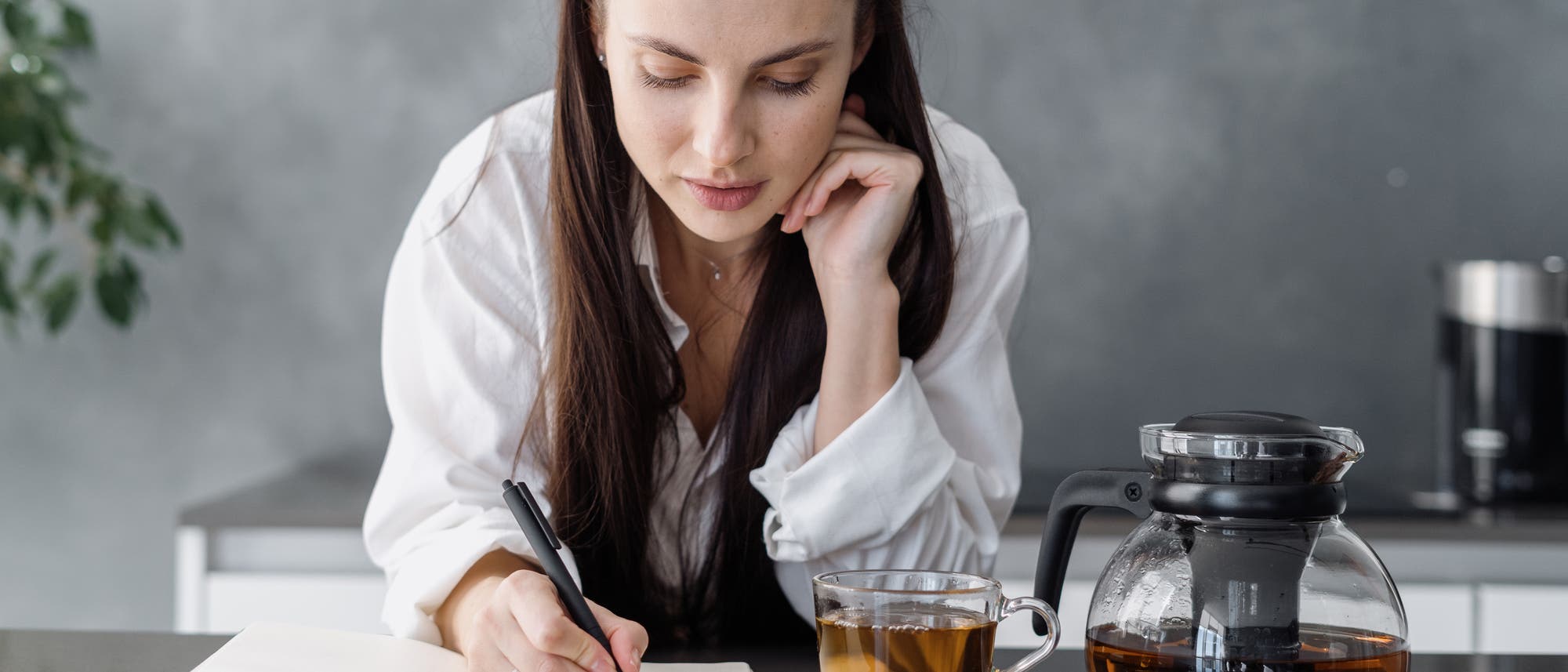 Junge Frau schreibt in ein Notizbuch, neben ihr eine Tasse und eine Kanne Tee.