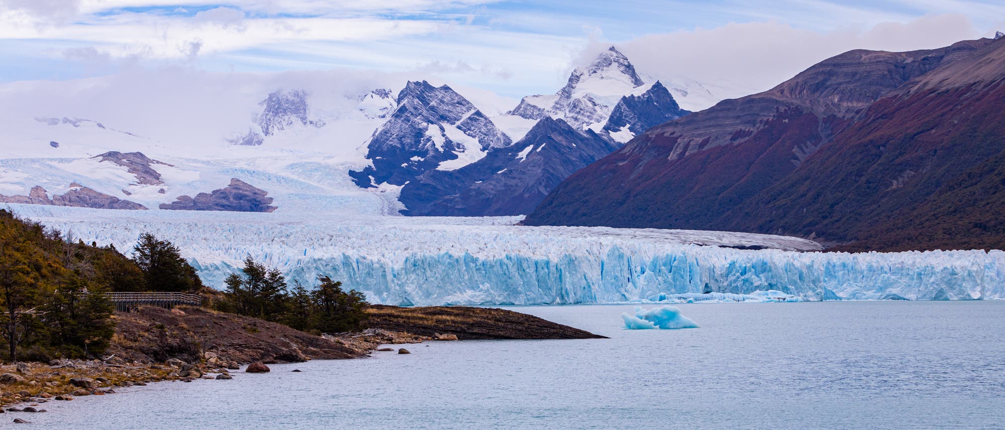 Die zerklüftete Zunge des Glaciar Perito Moreno in Patagonien mündet in einen See. Im Hintergrund ein schneebedecktes Gebirge.